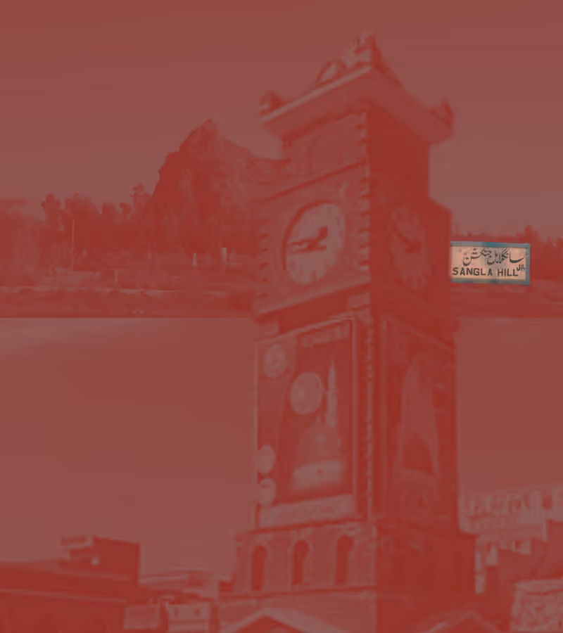 A red-tinted photograph of the clock tower in Sangla Hill, with the hill behind it and the Sangla Hill railway sign.